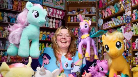 A woman in a room where there are shelves filled with different coloured toy horses. The woman has wavy blonde hair, glasses and she wears a blue jumper with images of the toy ponies on it.