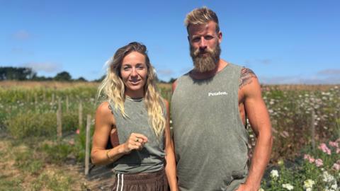 A woman with long blonde hair standing in a field next to a man wearing a vest, with a big beard and blonde short hair