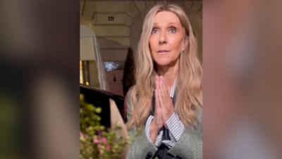 Celine Dion, in a grey suit, makes a thank you hand gesture when she is offered flowers by a fan.