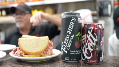 A sandwich and two types of cherry cola on a countertop in a deli.