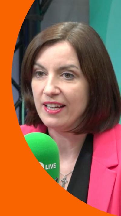 A woman with brown hair in a bob sits behind a mic. She is wearing a pink suit jacket over a black blouse.