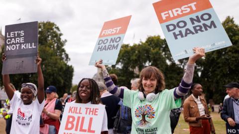 Anti-assisted dying campaigners react after MPs voted against the assisted dying bill. Three women stand in a line holding up placards which read: Let's Care Not Kill, Kill the Bill and First Do No Harm. They are smiling and wear t-shirts which support their campaign.