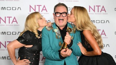 Paloma Faith in a black dress, Alan Carr in a turquoise suit and Amanda Holden in a black dress stand in front of a wall that says "NTA", with Alan smiling and holding a trophy and Paloma and Amanda kissing him on the cheek