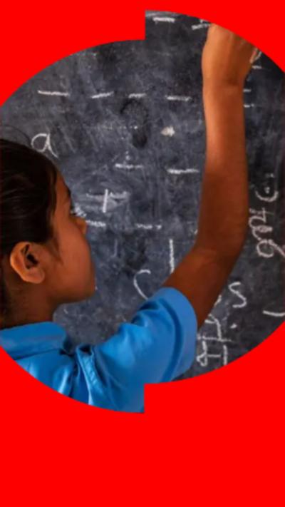 A young girl with dark hair and wearing a blue top, writes on a chalkboard.