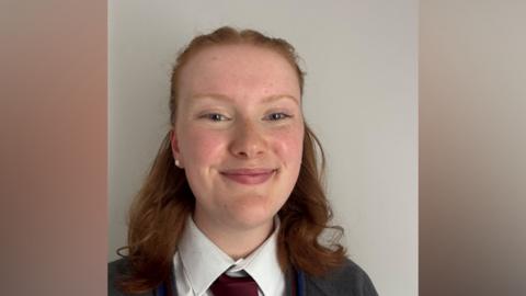 Connie is in her school uniform. Her auburn hair is tied back. She's standing against a wall. 