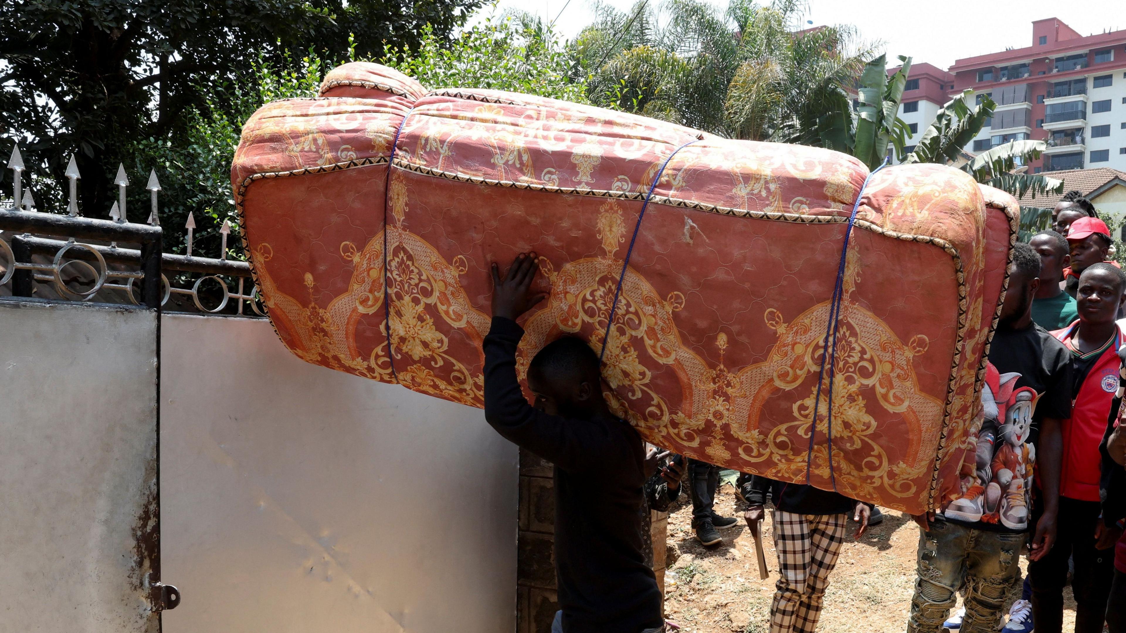 Man carrying red mattress over his shoulder. behind him is a line of people