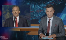 A man in a suit sits behind a talk show desk. In the top left is an image of another man talking on stage.