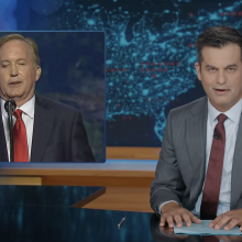 A man in a suit sits behind a talk show desk. In the top left is an image of another man talking on stage.