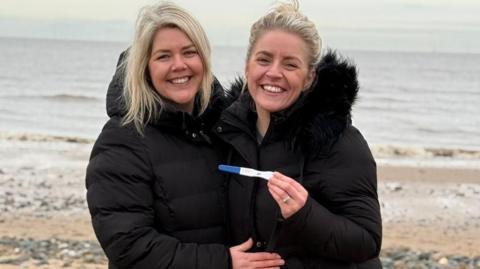 Two blonde, white women, one with her hair tied up and the other with her hair down, in big black coats, stand side by side with their arms around each other. Both are smiling and one of them is holding up a pregnancy test and her wedding ring is visible. They are stood on a beach and in the background there is sand, rocks, as well as the sea. 