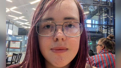 A selfie of a young woman with dyed red straight hair and glasses. She has a green sunflower lanyard round her neck.