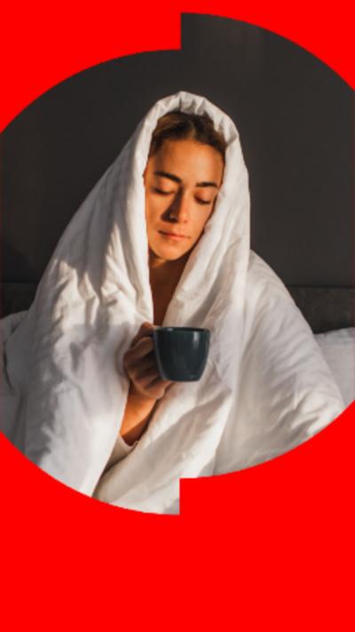 Young woman in bed with the duvet over her head, holding onto a mug.