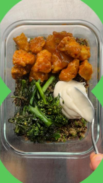 A glass food storage container filled with quinoa crispy tofu and broccoli. A spoon is ladling in yoghurt from the side.
