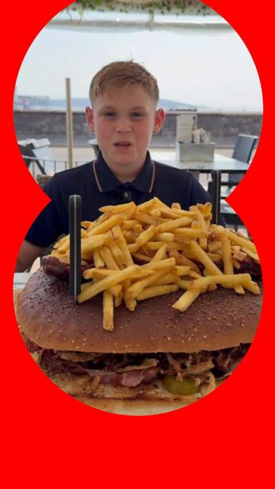 A young boy sitting in front of an enormous burger with chips on top of it. 