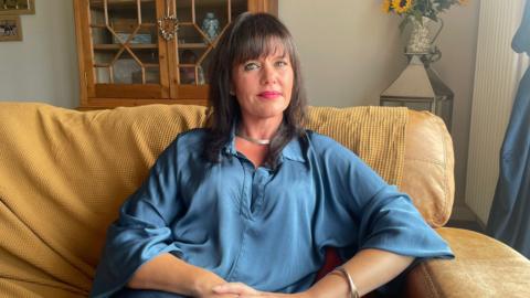 A woman with dark brown hair and a fringe is sat on a mustard-colour sofa wearing a blue silk shirt. She has her hands on her lap.