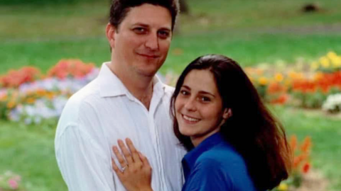 John Resta is on the left, he has dark hair and is wearing a white linen shirt and his wife Sylvia is on the right, with dark long hair and a blue short-sleeved shirt. She is pressed against him, her left hand on his chest. It has a wedding ring on it. They are both smiling. Behind them is grass with flowers.