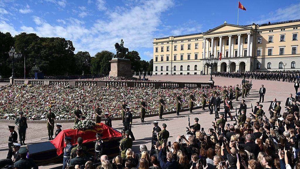 Crowds of people are seen lining the road behind a barrier outside the royal palace in Oslo as King Harald's coffin, draped in a flag, is pulled on a stretcher 