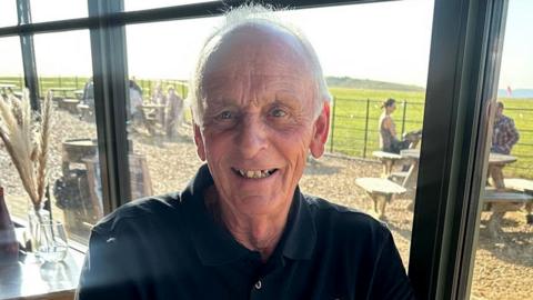 An older man wearing a black polo shirt, sits at a restaurant table with views of the countryside. He is smiling at the camera.  