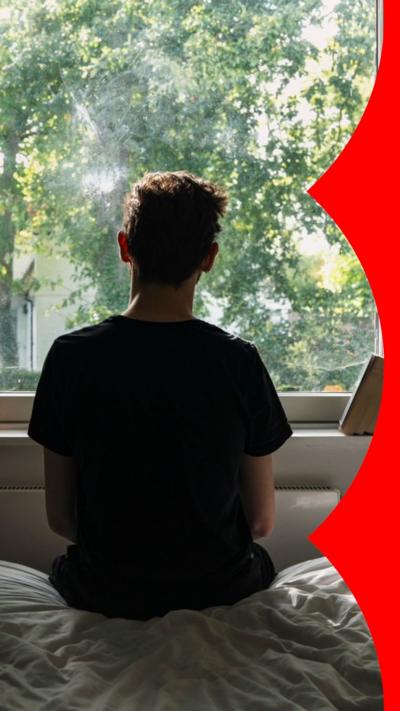 A young person sits on a bed in front of a window