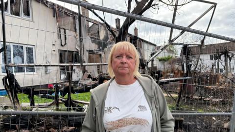 A middle-aged woman with shoulder-length blonde hair stands in front of a row of fire damaged houses which are fenced off.