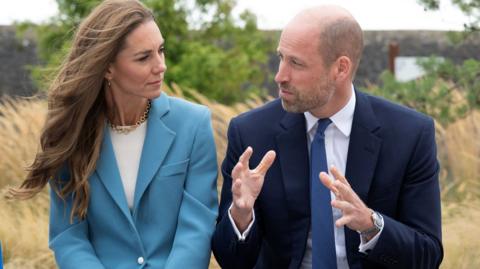 Catherine, Princess of Wales and Prince William look at each other. She is wering a teal suit and he a dark blue one with his hands strateched out in front of him