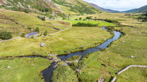 A green valley in Eryri national park. It has steep sides and is dotted with trees, a winding stream and a small stone cottage