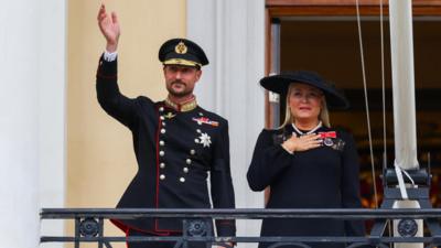 King Haakon waves to the crowd from the balcony of the royal palace, as Queen Mette-Marit stands next to him