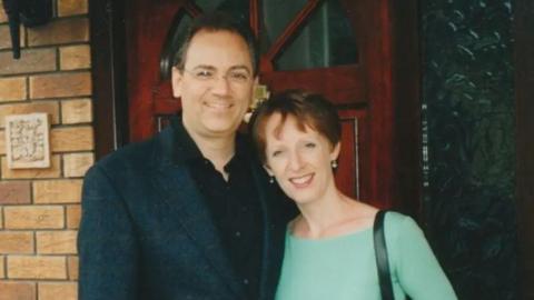 A picture of Charles Wolf and his wife smiling at the camera. They are stood outside a door to a house. He is wearing a dark suit jacket and a black shirt. He has short, brown hair and is wearing spectacles. She is leaning into him, wearing a green jumper. She has short, red hair.