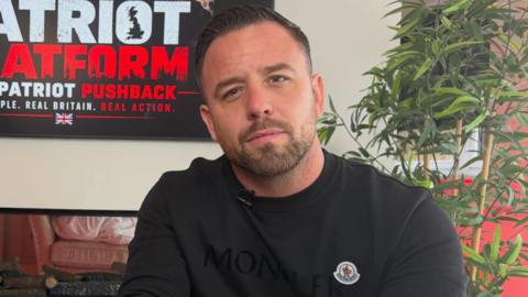Danny Tommo wearing a black t-shirts with clip-on microphone looks into camera with a poster saying "Patriot Platform the patriot pushback" behind them