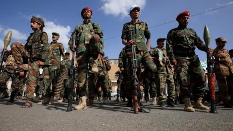 A dozen Houthi men wearing combat clothes, berets and holding large guns. The sky is blue and the men are looking straight ahead. 