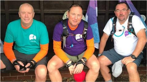 Three men sit side by side. The man in the centre and the man to the right are carrying backpacks. The man to the left has short grey hair and is wearing a green T-shirt, an orange long sleeved top underneath and grey shorts. The man in the centre has short brown hair, the same orange top, with a purple T-shirt over and khaki shorts. The man to the right has short dark hair and a beard and glasses. He is in a white T-shirt and grey shorts.