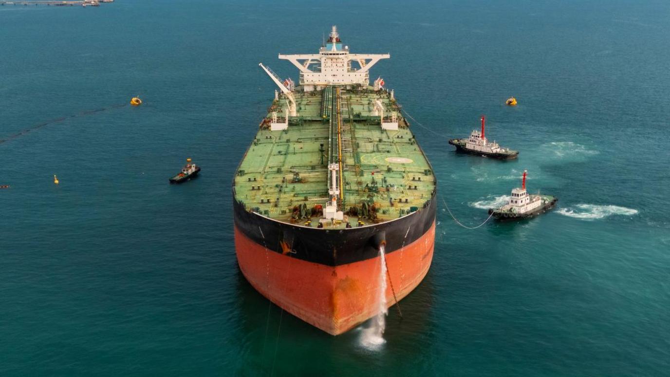 Stock image of an oil tanker pictured front on, with several tugs surrounding the ship