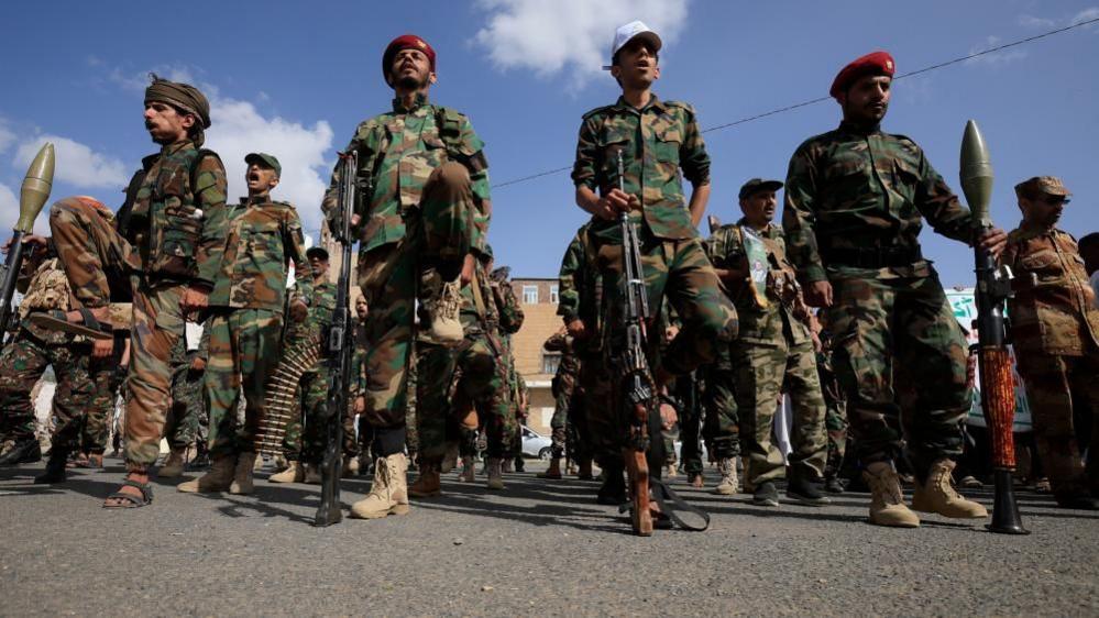 A dozen Houthi men wearing combat clothes, berets and holding large guns. The sky is blue and the men are looking straight ahead. 