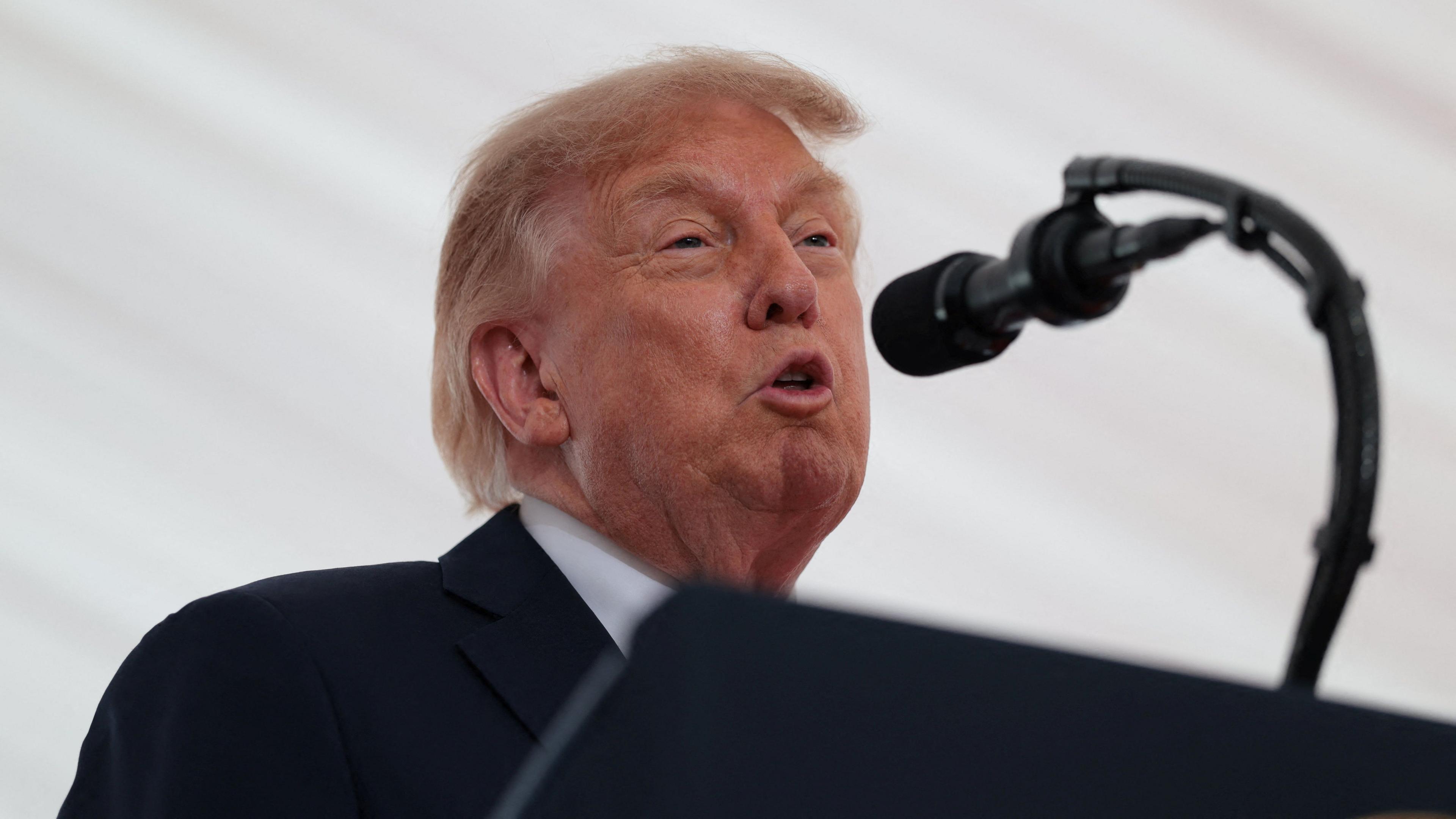 Trump is speaking into a mic. He has a black suit on with a white shirt. A white backdrop is behind him.