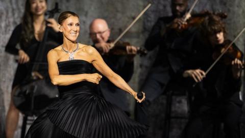 Dion dancing in a black dress, holding a microphone, while musicians play behind her