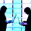Two men stand silhouetted against an illuminated blue and purple background. They are facing each other, but looking down at laptops they are holding in their hands. 