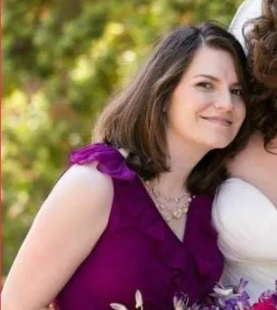 A woman with dark hair, wearing a purple dress and necklace, smiles at the camera.