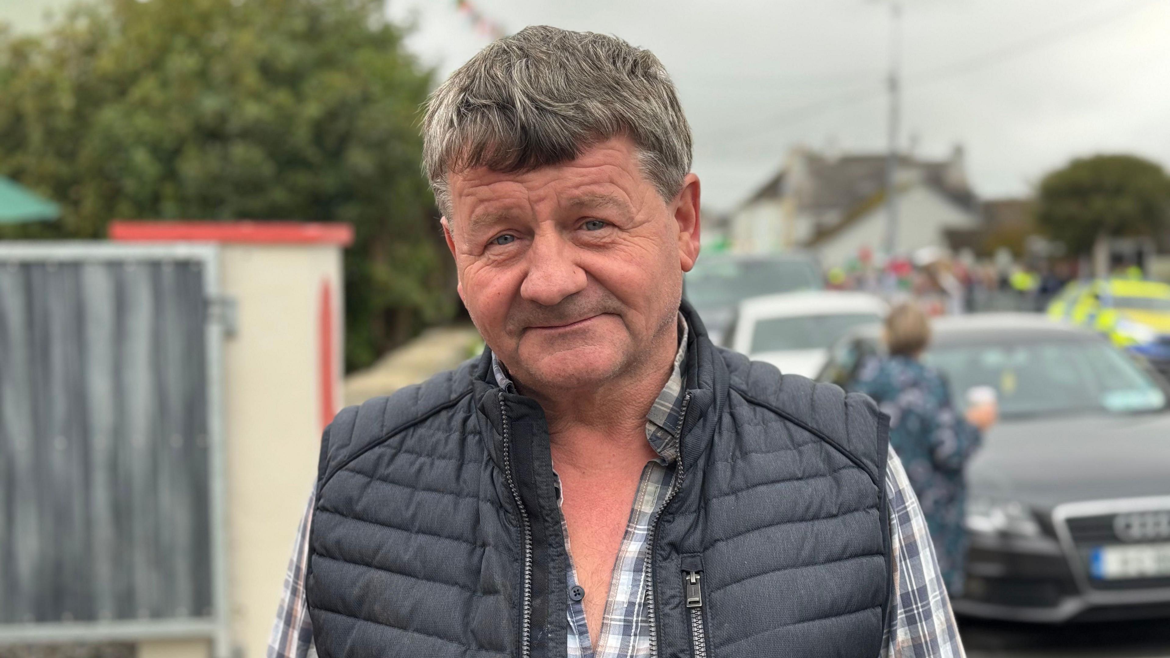 A man is standing on the street. He's looking into the camera. He has a checked shirt on with a vest jacket on. A police car is out of focus in the background. 