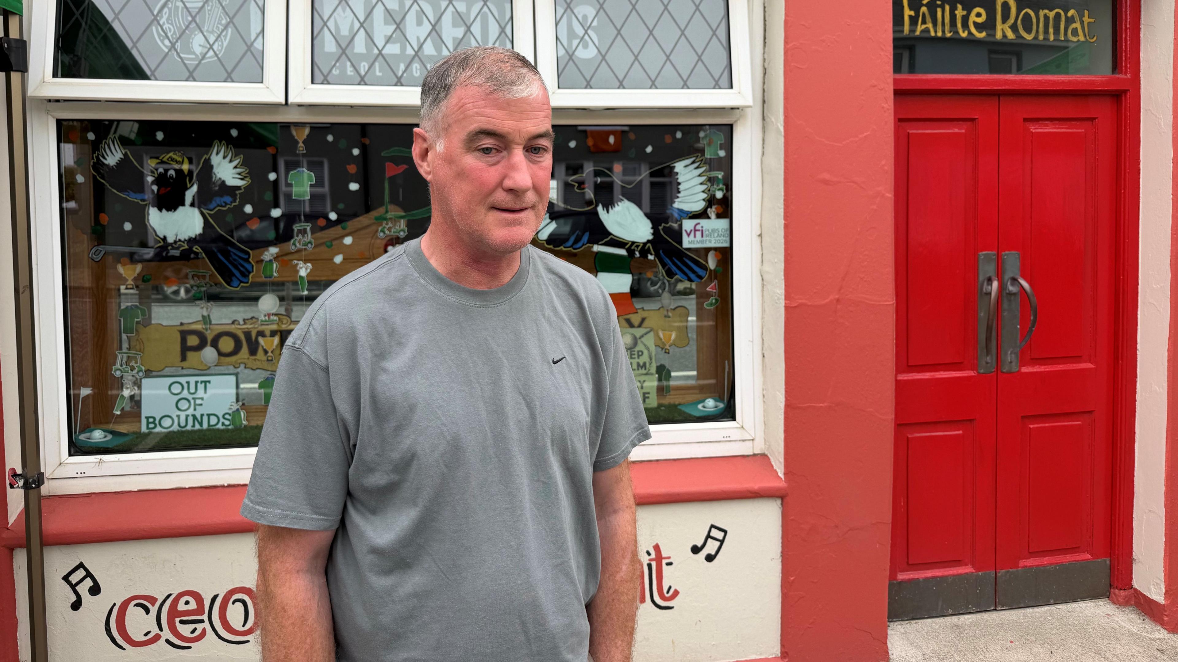 Seoirse Comerford is standing outside a bar. He's wearing a grey top. The door to the bar is red and says failte romat over it. There are bird designs on the window. 