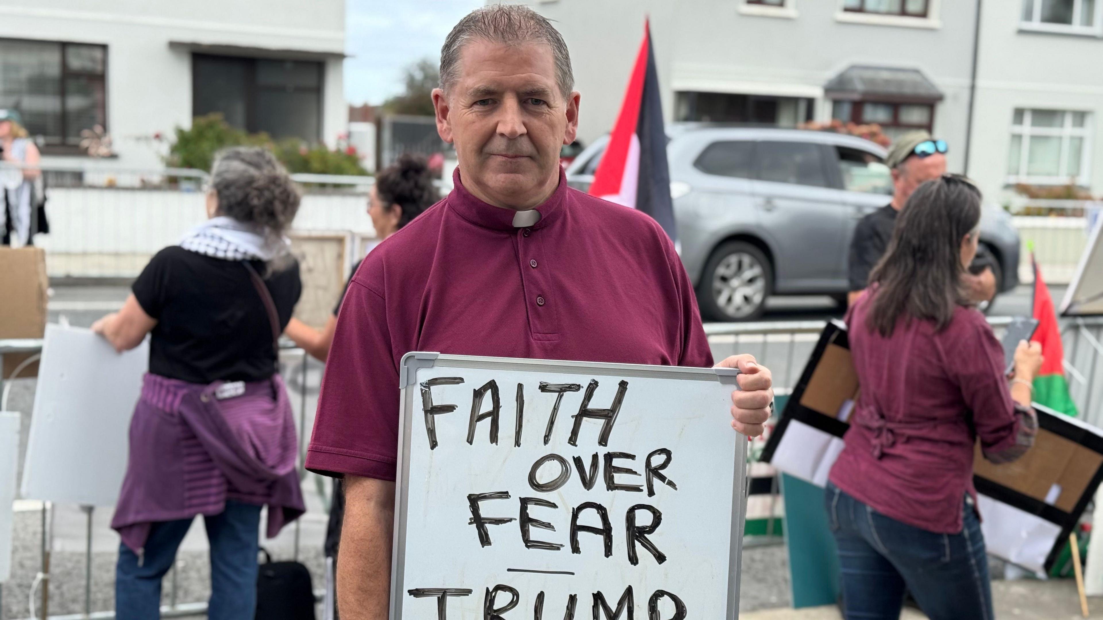 A man wearing a white collar on his maroon top is holding a white board saying faith over fear. Trump get out of here. 