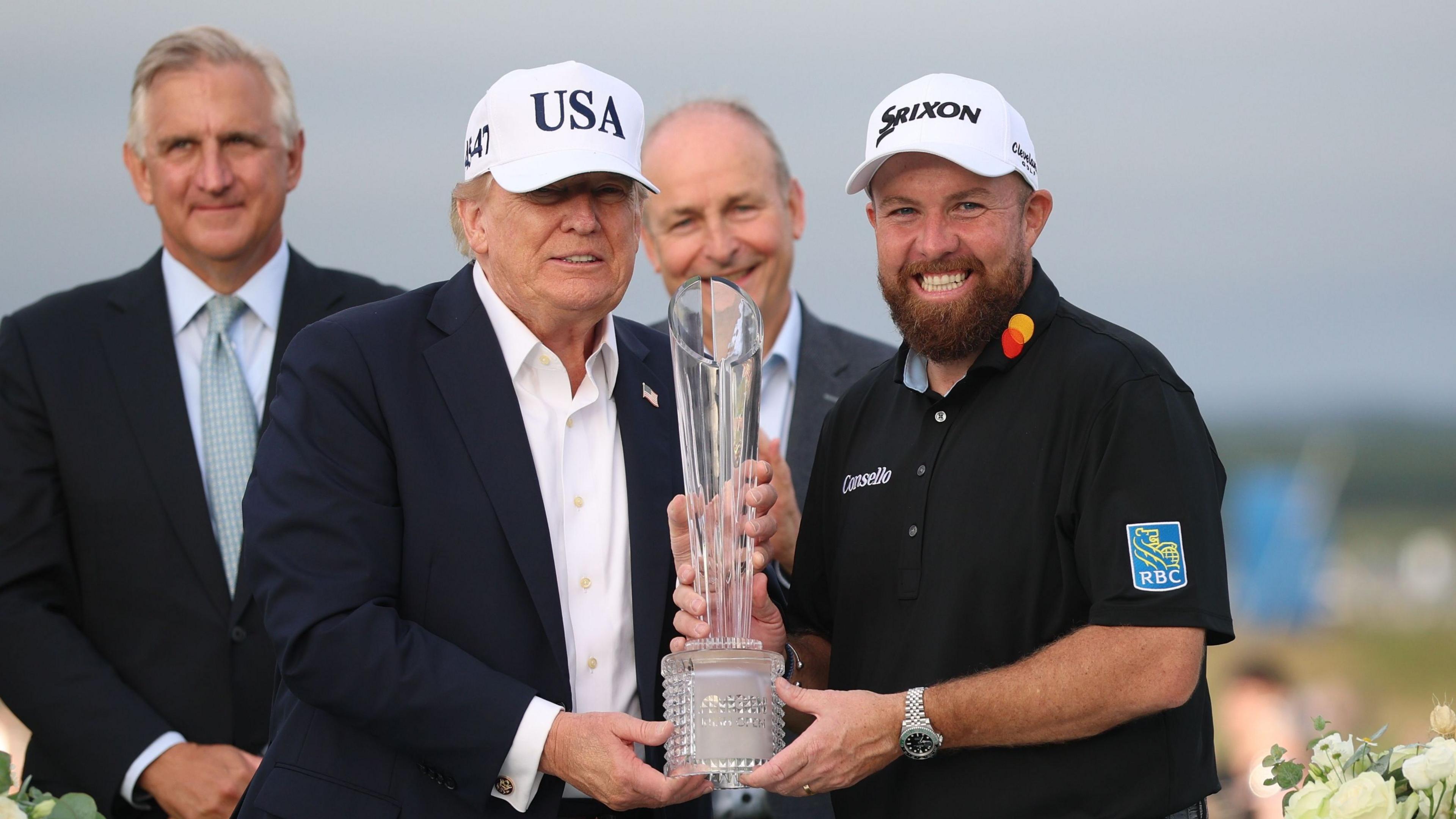 Two men stood, holding a trophy. They are both smiling. On the left is Donald Trump, wearing a navy suit and a white hat with 'USA' written on it. On the right is Shane Lowry, wearing a black polo shirt and a white hat. Behind them stand two men. 