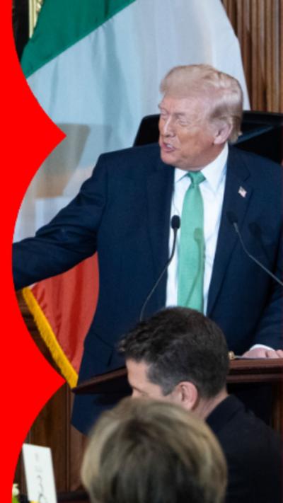 Donald Trump wearing a green tie, stood on stage in front of a room full of people, with the Irish flag and the American flag behind him.