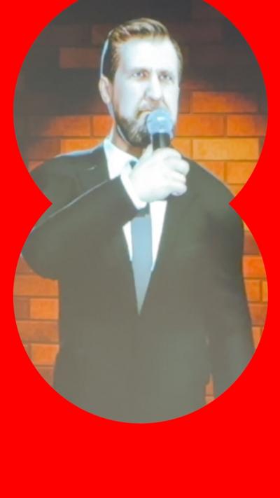 A screen showing the AI avatar of comedian Garrett Millerick. It is wearing a black suit, white shirt and blue tie. it has short dark hair, a beard and moustache.