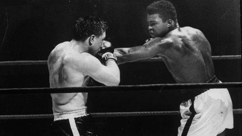Black and white photo of Muhammad Ali and George Chuvalo boxing in a ring. Chuvalo, in dark shorts with a white stripe, has his arms up, blocking a punch thrown by Ali, who is in white shorts with a dark stripe. 