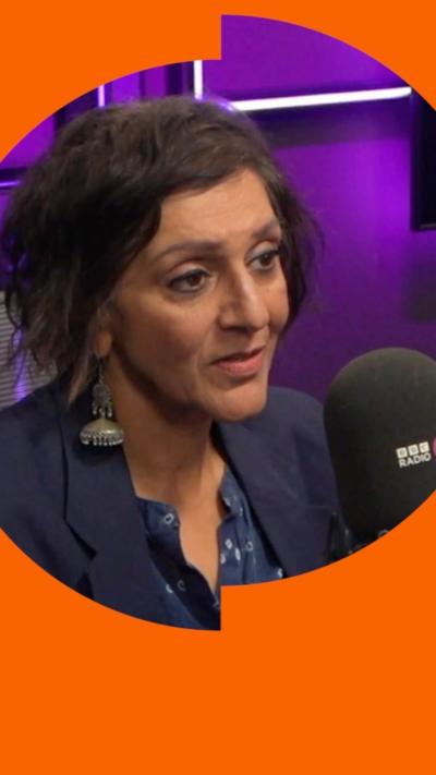Meera Syal is sitting with a microphone in front of her in a purple radio studio. She is wearing a blue blazer and top and has short brown hair.