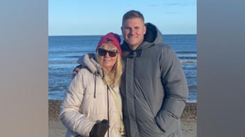 A blonde woman in a pink hat, white winter coat and sunglasses stands with a taller man in a grey winter coat. The man in the grey coat has his arm around the woman.  Behind them is sand and the sea.
