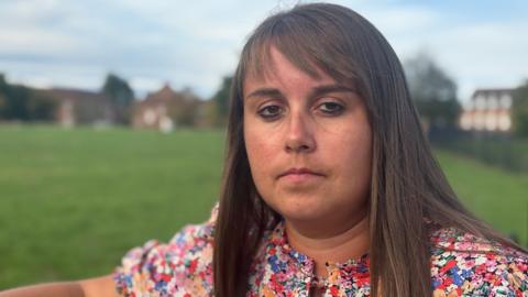 Becky Jones has shoulder-length brown hair with a fringe. She is standing in a park and is wearing a dress with multi-coloured flowers on it. 