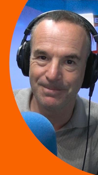 Martin Lewis is wearing headphones and sitting behind a microphone. He has short brown hair and wears a grey T-shirt.