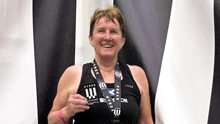 Carole Munro smiles at the camera wearing a medal and holding a patch. She is standing in front of a black and white striped backdrop and is wearing a black vest top.