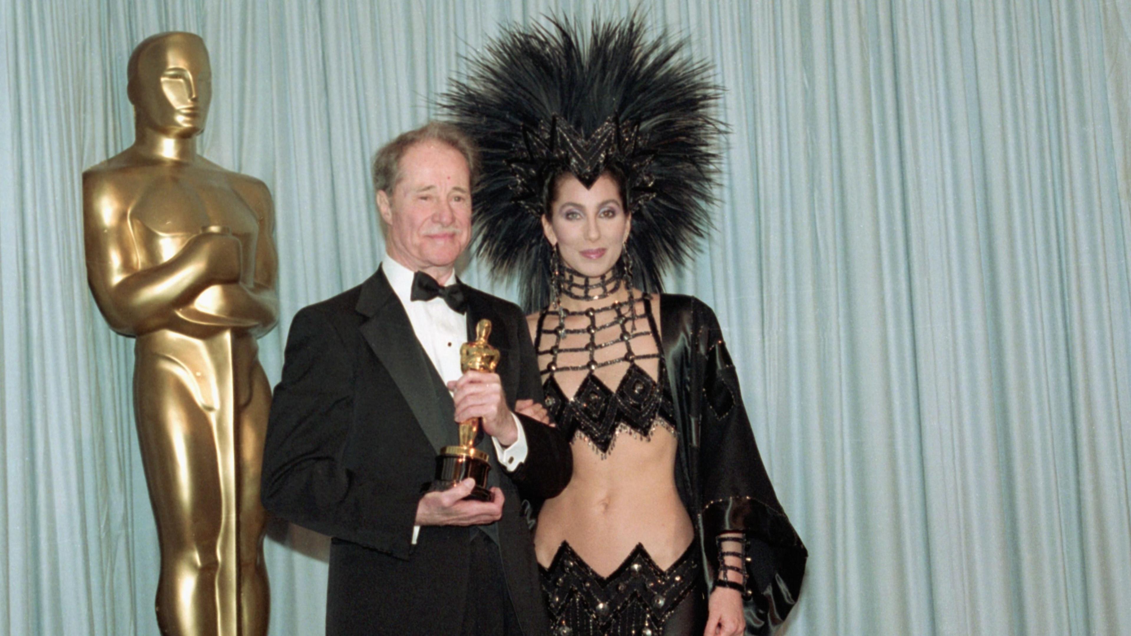 Cher's outfit is a black bejewelled skirt and top that shows all of her stomach, and she has a huge black headpiece on that has many black feathers sticking into the air