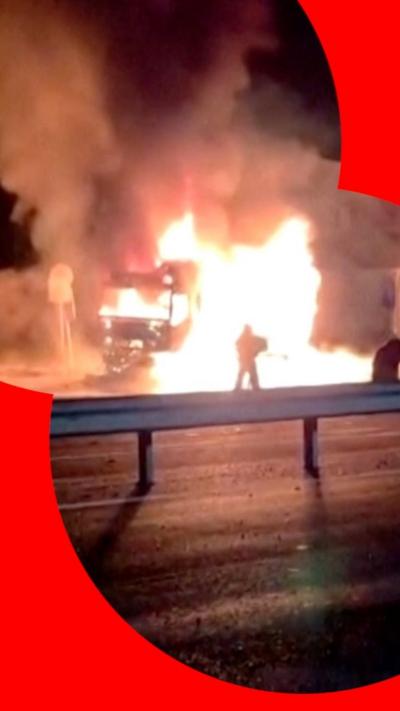 A petrol station is on fire at night, with large flames rising up into the sky. It is in the background of the picture. In the foreground, there is a large road with a metal barrier in the middle.